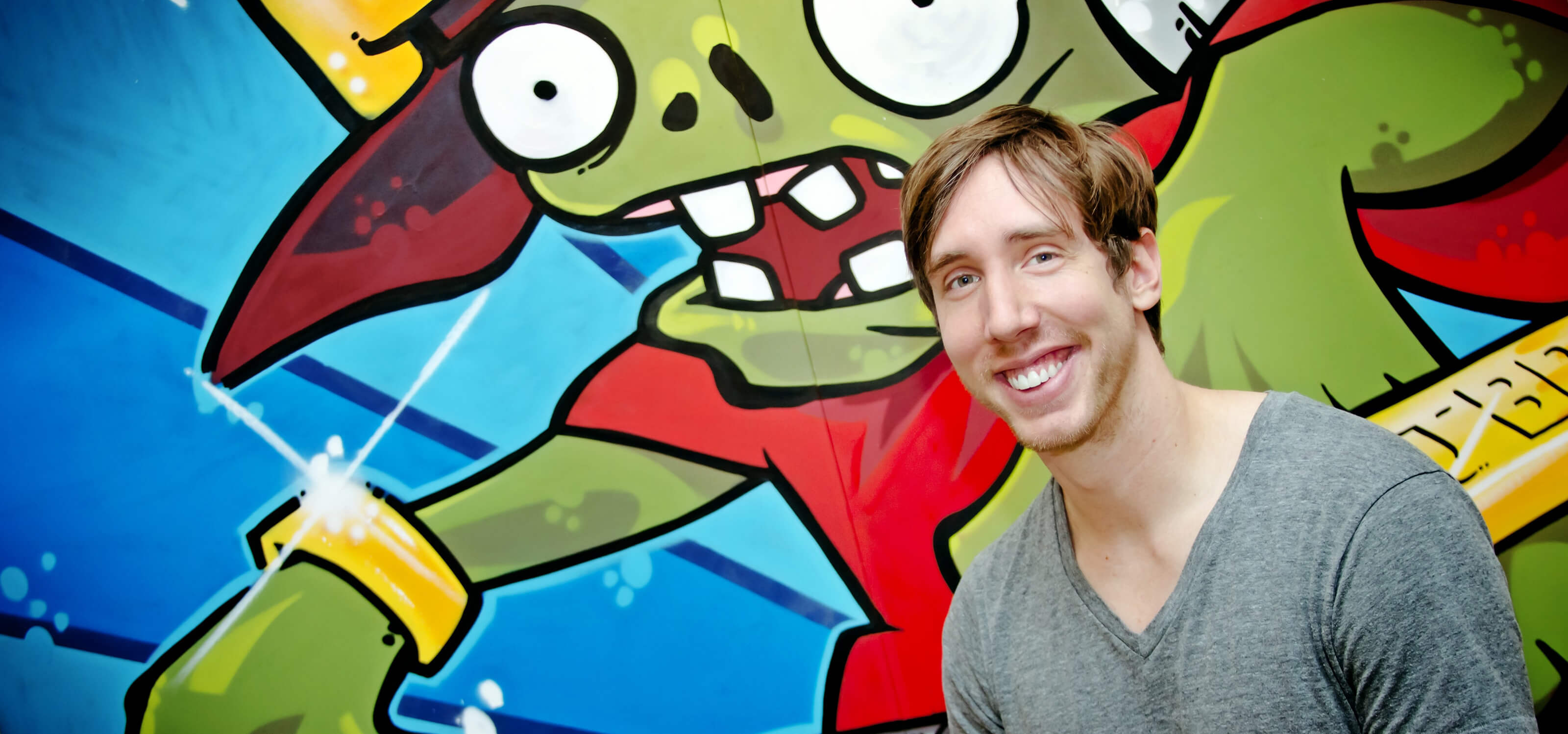 DigiPen BFA alumnus Mark Barrett smiling in front of large grafffiti-style wall painting of a zombie at PopCap's office