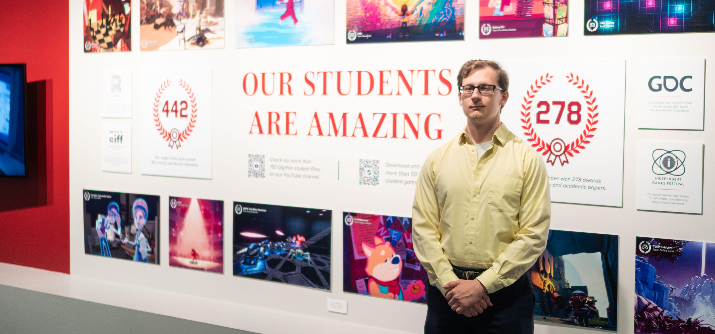 Joseph Crump poses with his hands folded in front of a DigiPen campus hallway display.