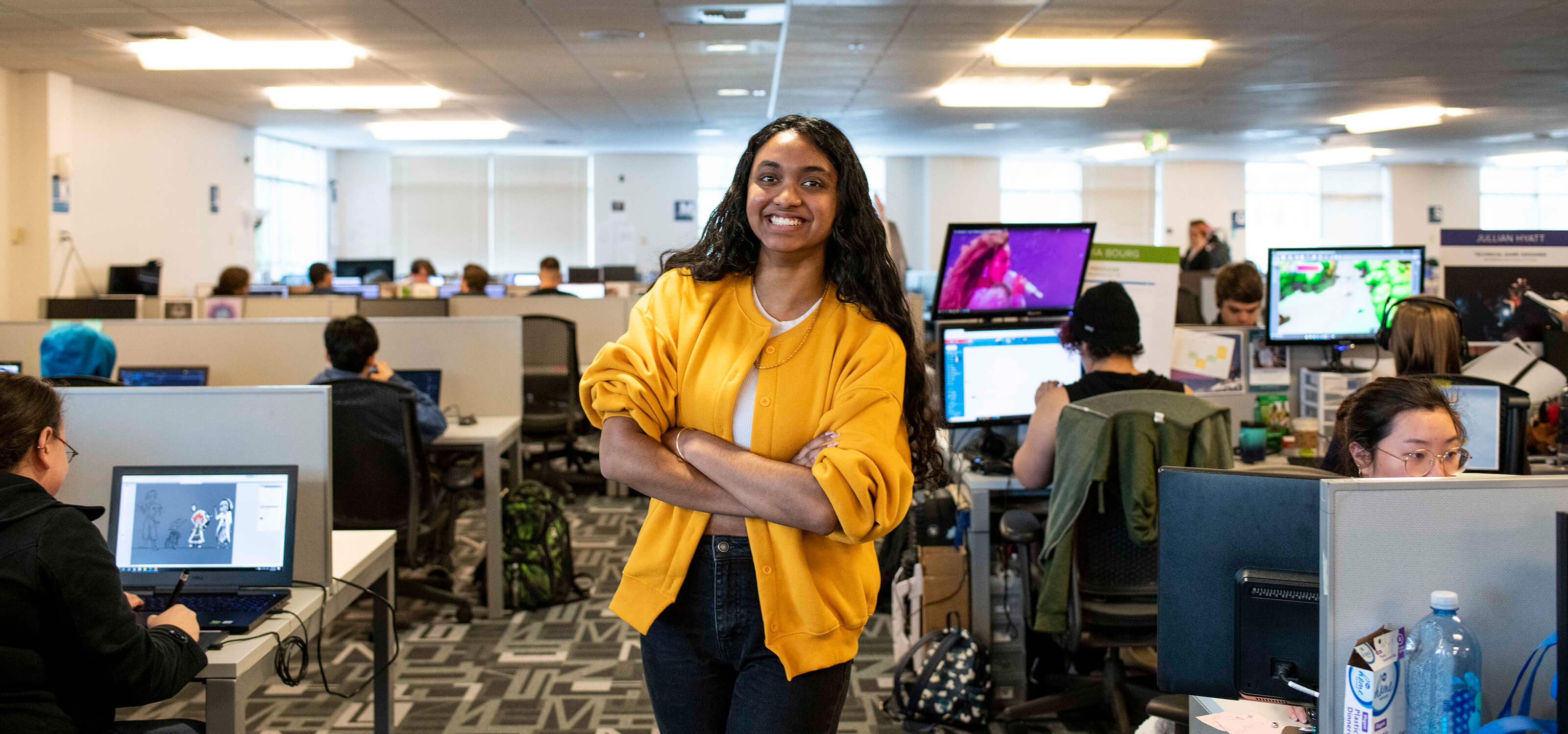 Student Shannon Parayil smiles and stands in the Tesla computer lab on campus, in between rows of team spaces where students are working on projects.