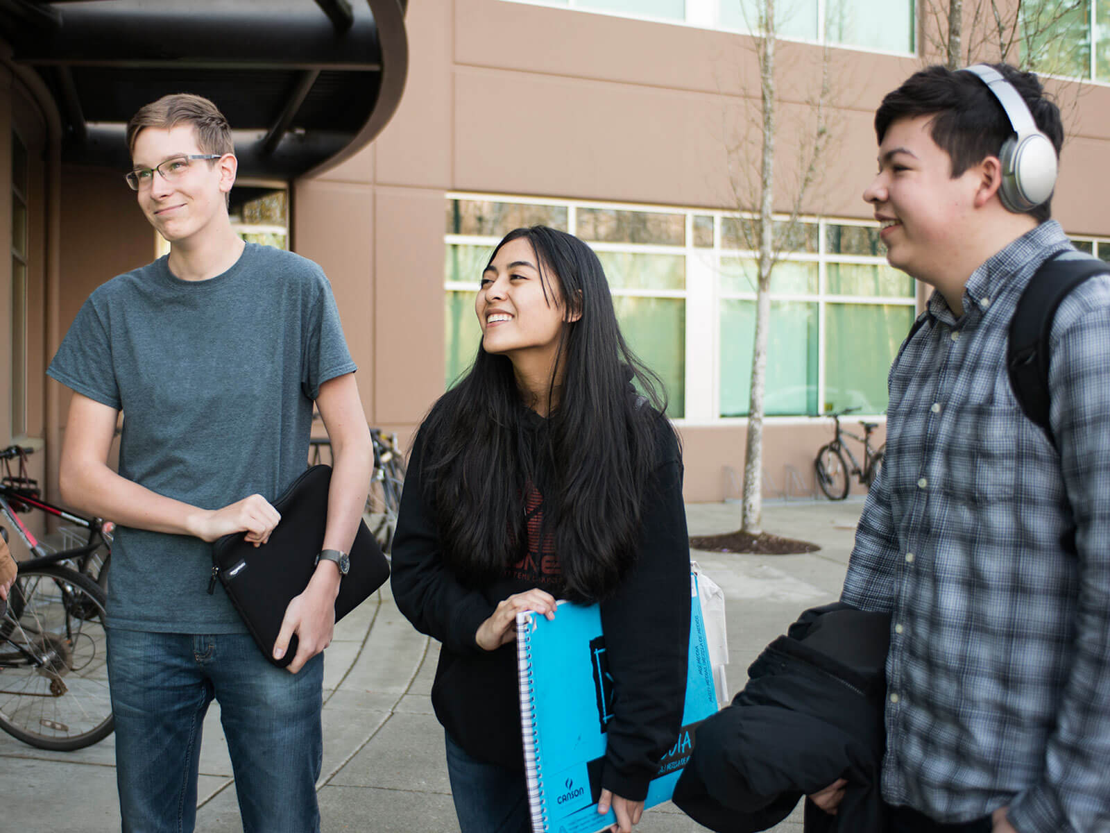 A group of students stand smiling at each other in front of the first-floor lobby of a brown building.