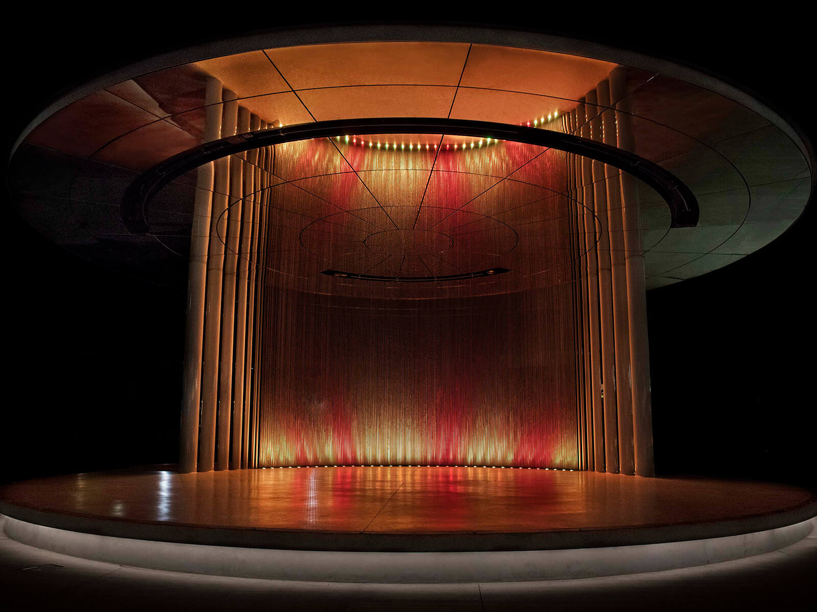 A circular structure with 380 etched metal rods lit in shades of red and yellow at night