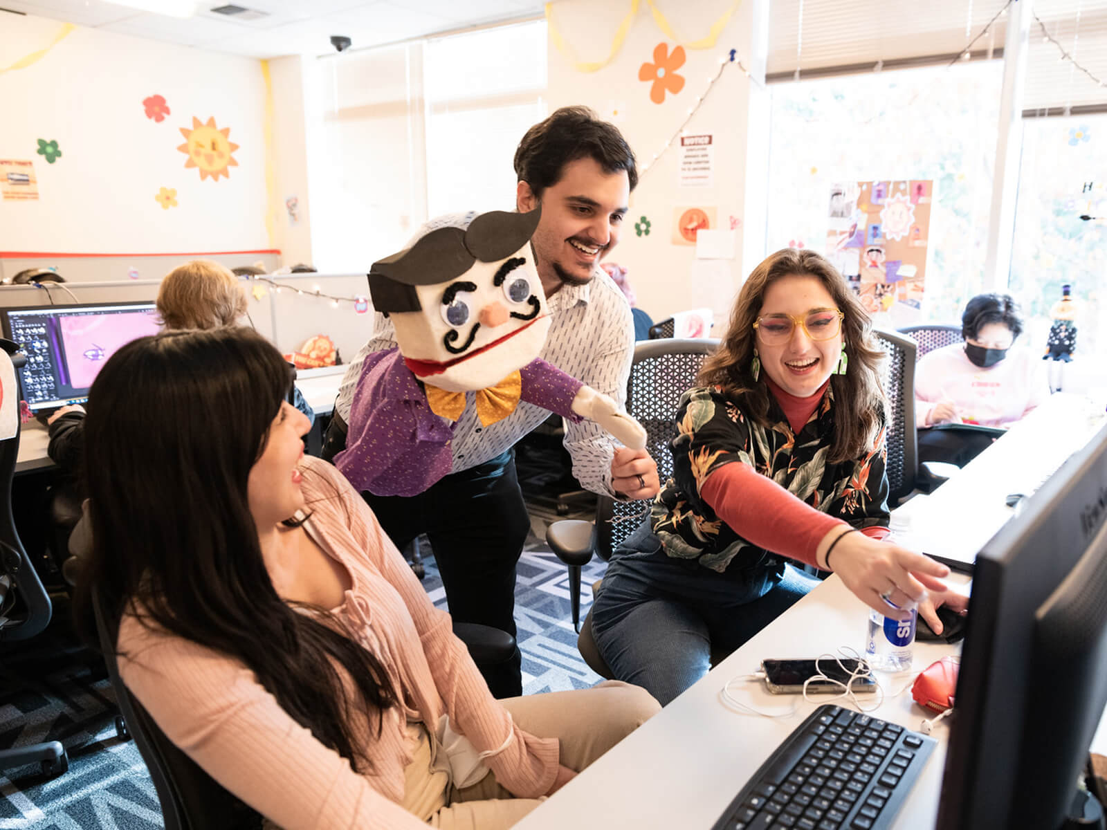 Three DigiPen students, one holding a puppet, smile as they look at a computer in the campus production lab.