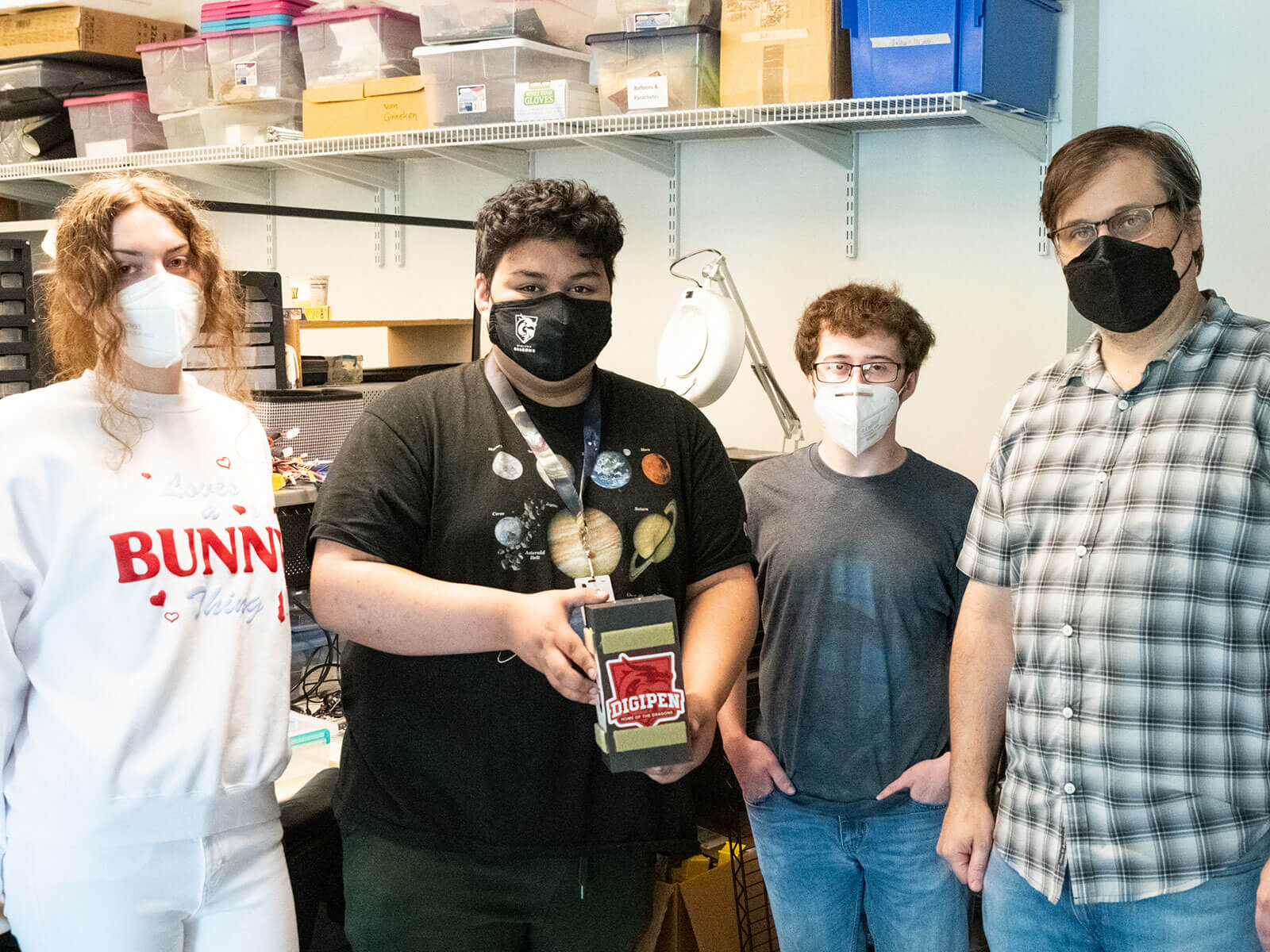 Three students and one professor stand together with student in center holding small payload box.