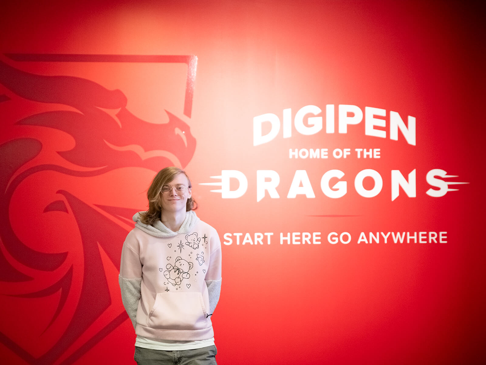 Student Tetra Chaney poses in front of a red wall with DigiPen’s Dragon logo on it and the words, “DigiPen Home of the Dragons — Start Here Go Anywhere.”