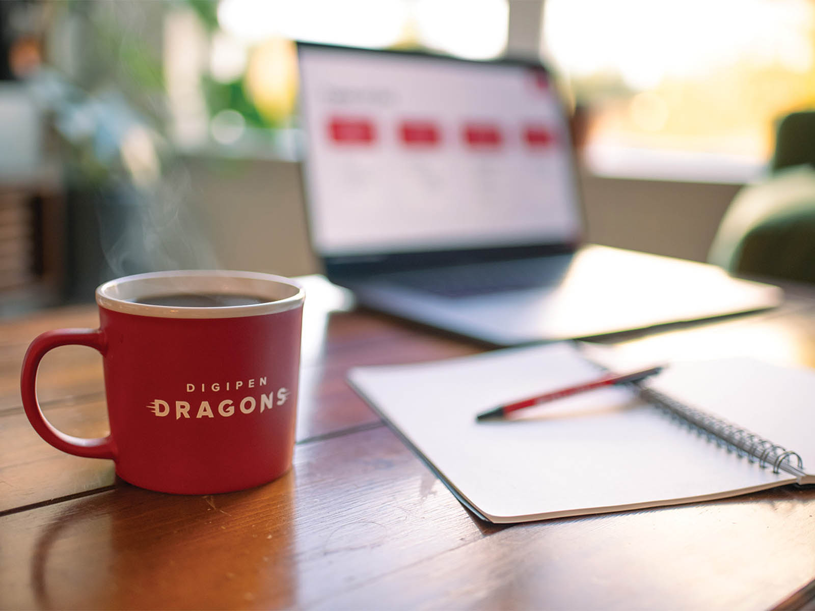 A filled DigiPen branded coffee cup sits next to an open notebook and laptop