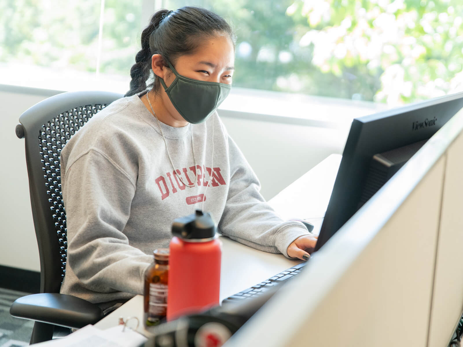 A student wearing a facemask and DigiPen sweatshirt sits in front of a computer screen.