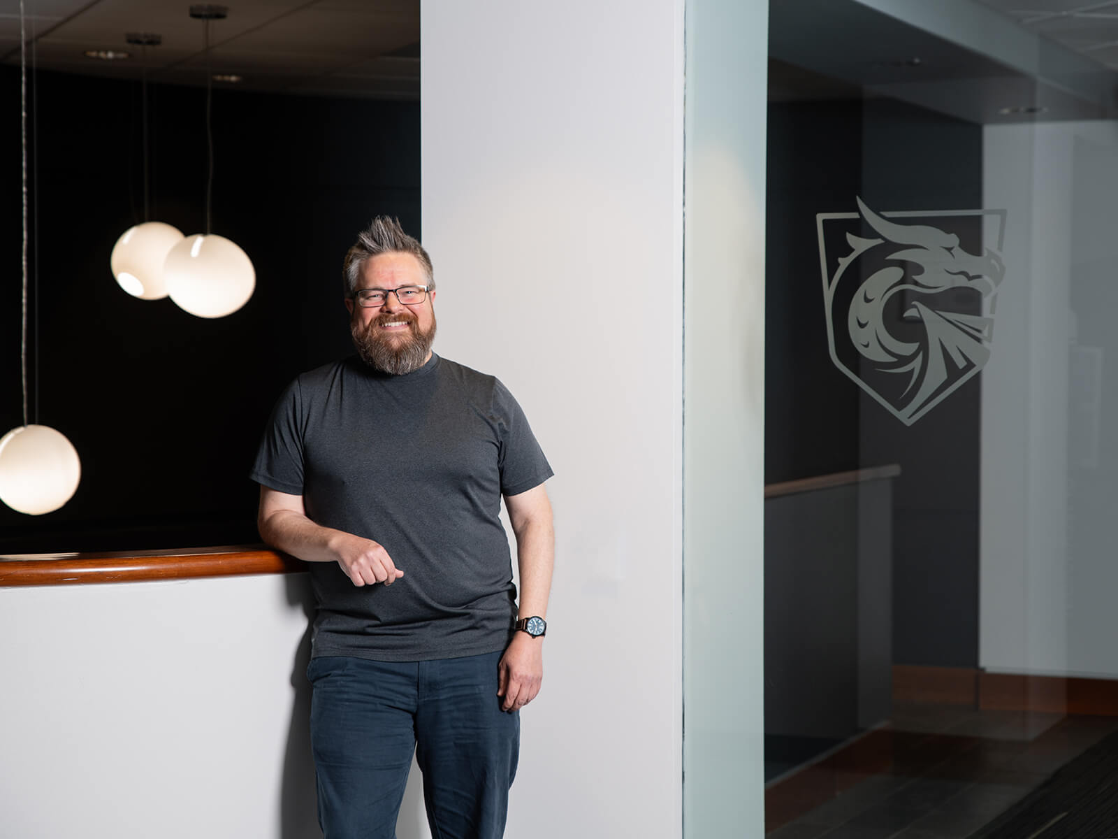 Chris Poplin smiles on campus standing next to a DigiPen Dragons logo decal on glass.