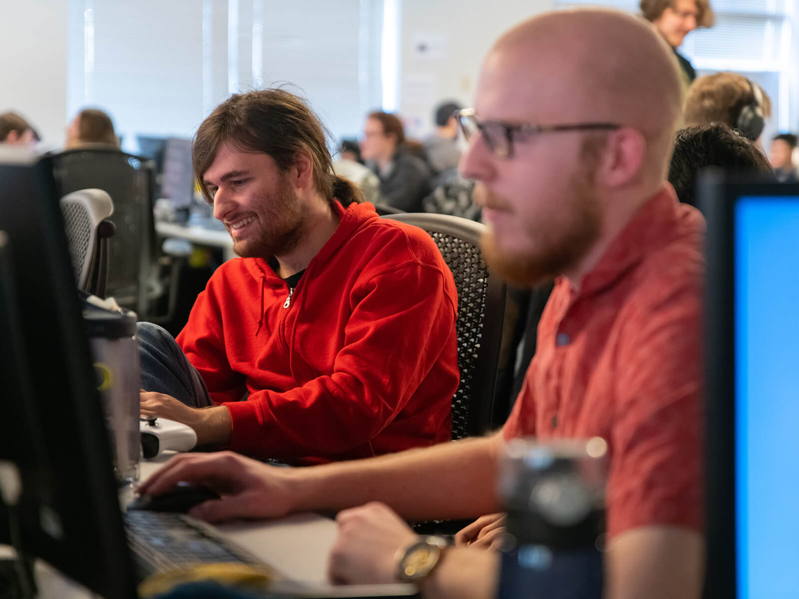 A DigiPen student in a red hoodie smiles as he works on his game project in the Tesla production lab.