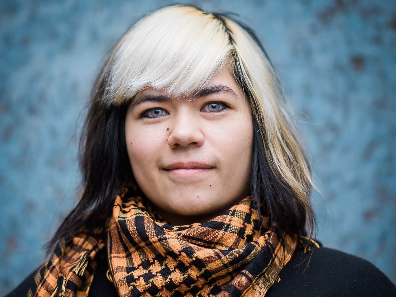 A headshot of Idris Grey MacChruiteir wearing an orange scarf on a blue background. 