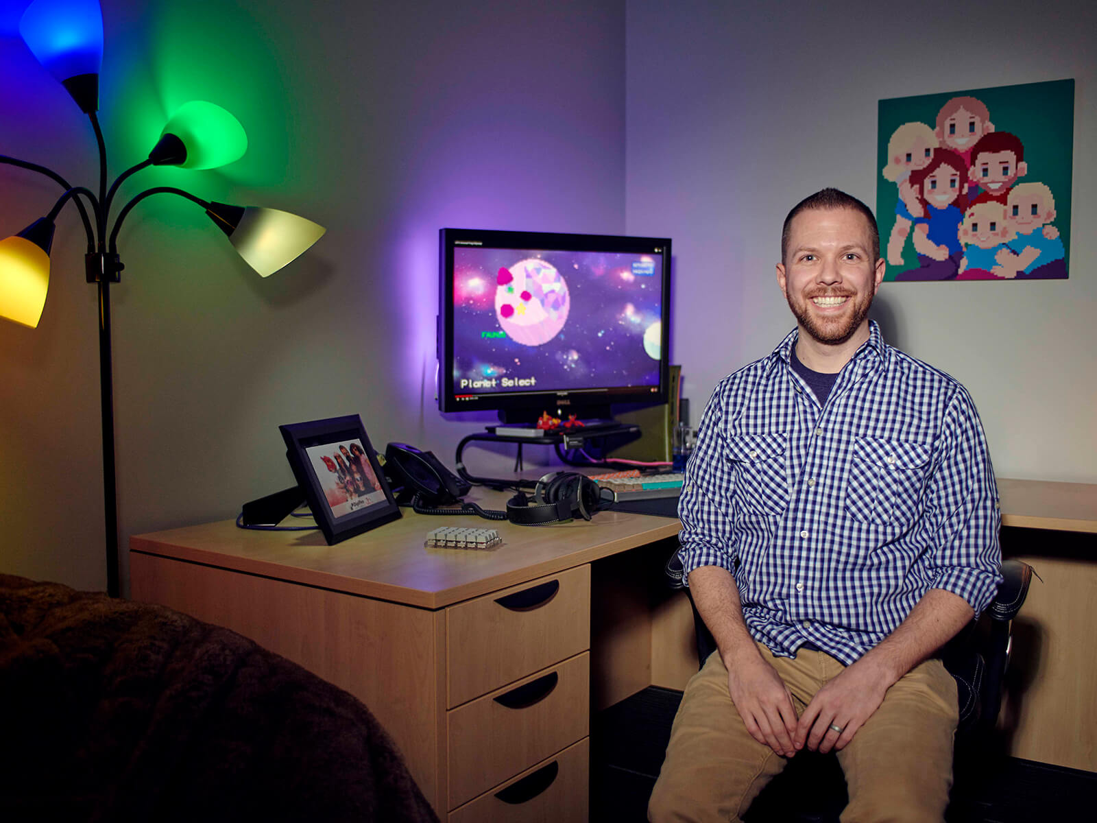 Justin Chambers smiles facing camera at his office desk, dark room decorated with objects and illuminated by colorful lamps.