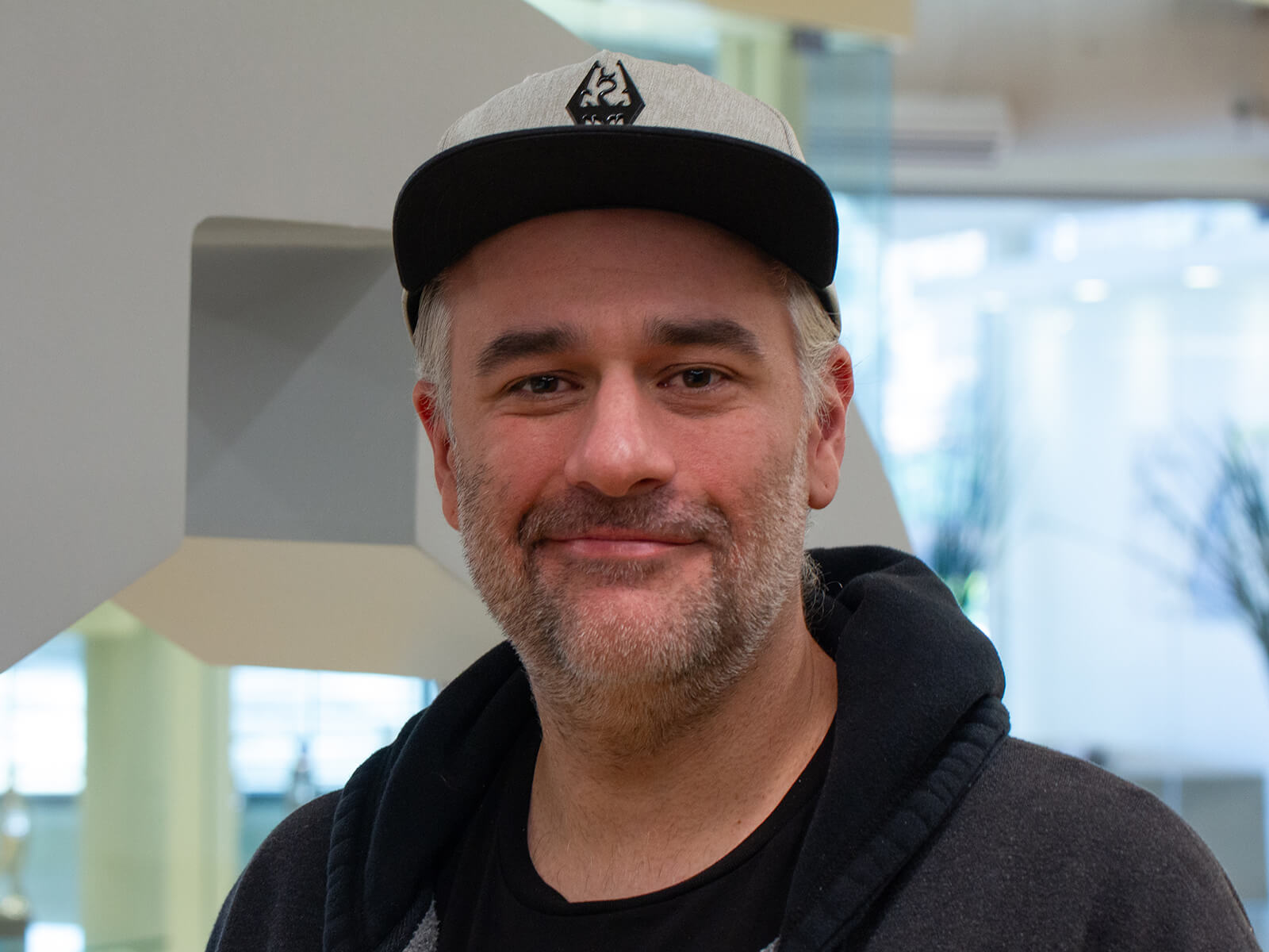 DigiPen alum Anthony Saulls smiles for the camera wearing a hoodie and a baseball hat with the Skyrim logo on it.