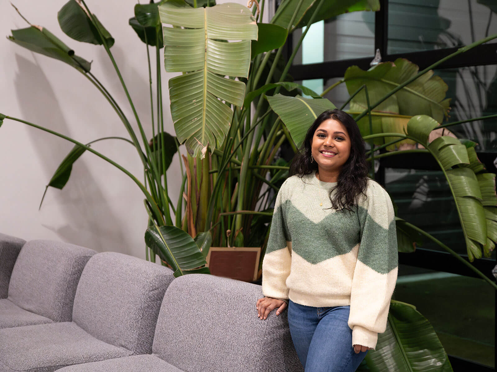 DigiPen graduate Neha Chintala smiles standing next to a couch and some large, leafy plants.