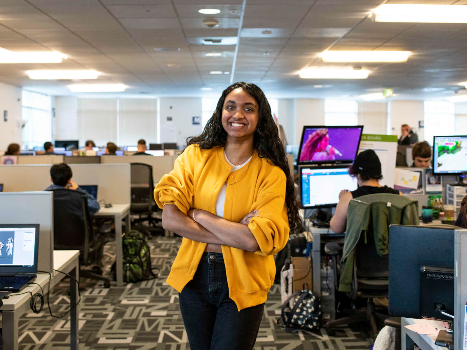 Student Shannon Parayil smiles and stands in the Tesla computer lab on campus, in between rows of team spaces where students are working on projects.