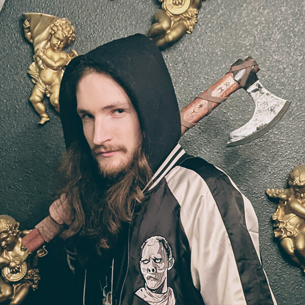 Devin Brom wears a black hood and holds an axe over his shoulder in front of a black wall with gold cherubs on it.