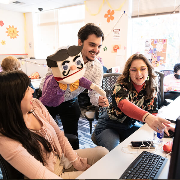 Three DigiPen students, one holding a puppet, smile as they look at a computer in the campus production lab.