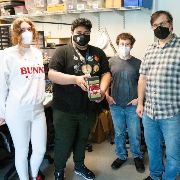 Three students and one professor stand together with student in center holding small payload box.