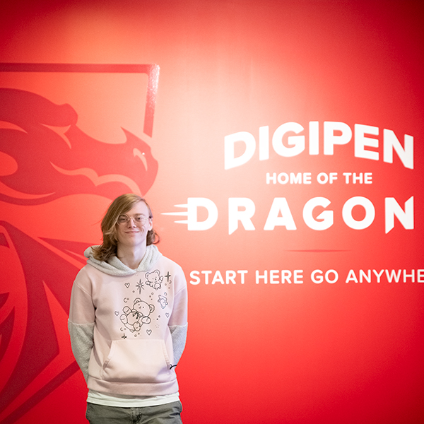 Student Tetra Chaney poses in front of a red wall with DigiPen’s Dragon logo on it and the words, “DigiPen Home of the Dragons — Start Here Go Anywhere.”