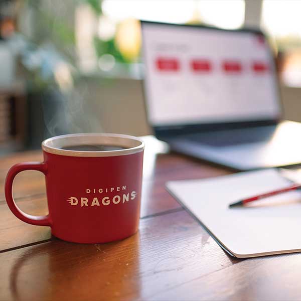 A filled DigiPen branded coffee cup sits next to an open notebook and laptop