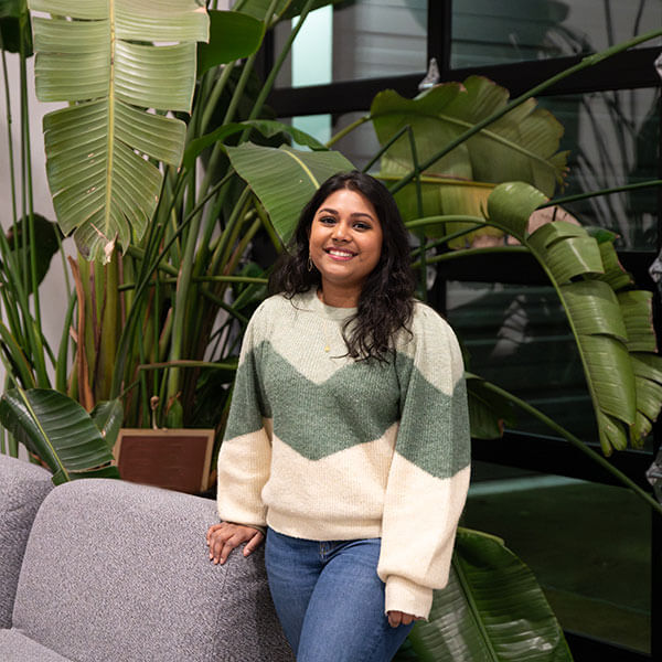 DigiPen graduate Neha Chintala smiles standing next to a couch and some large, leafy plants.