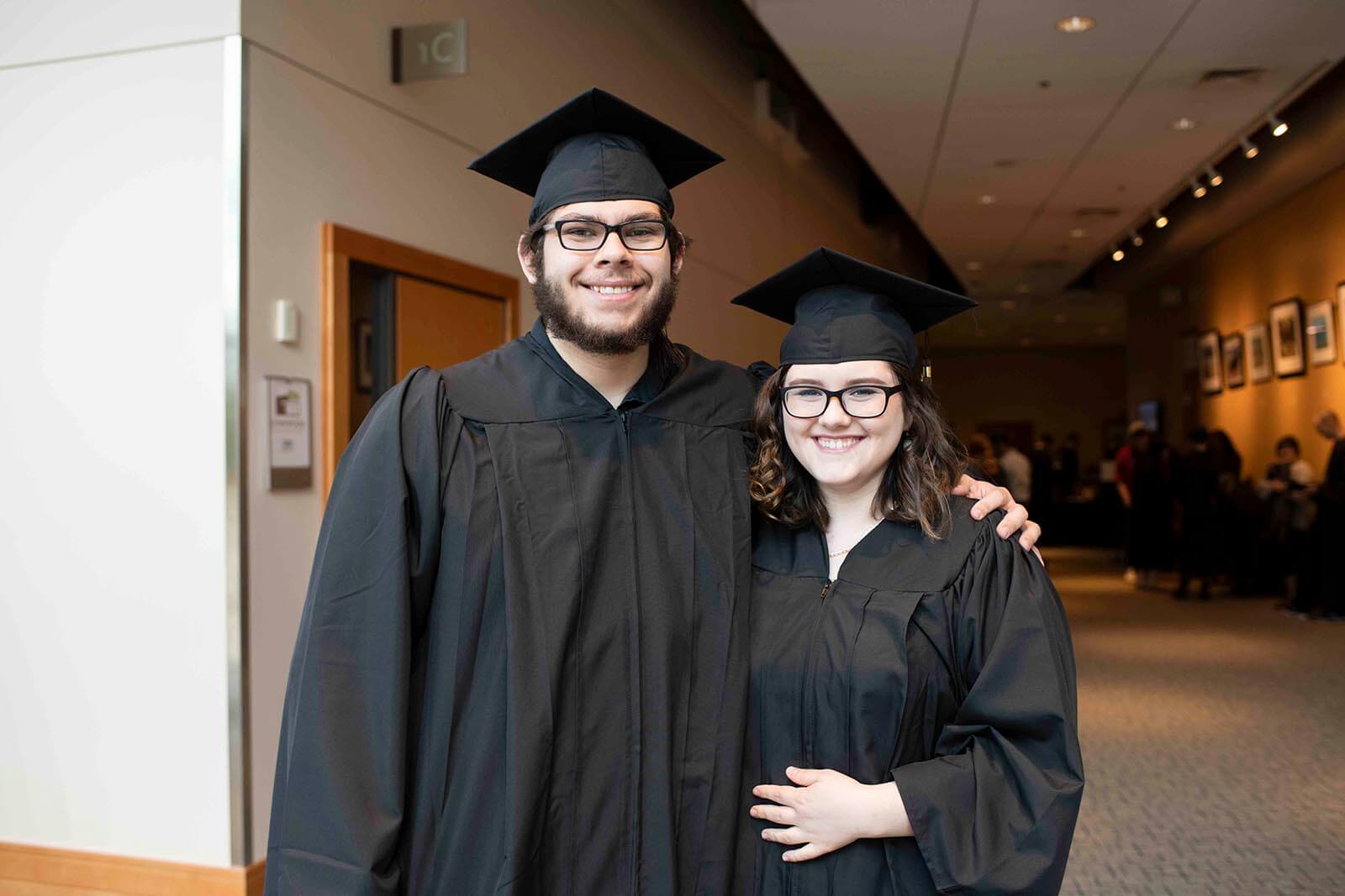 A man and woman in graduation robes and mortarboards pose for a photo in a convention center hallway.