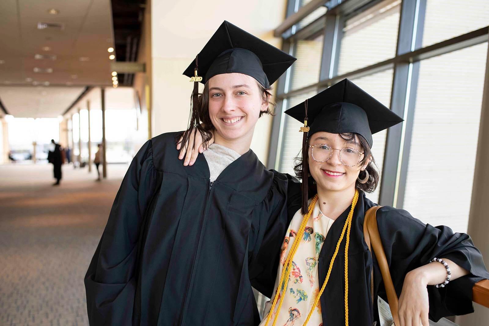 Two students in graduation robes and mortarboards pose for a photo in a convention center hallway.