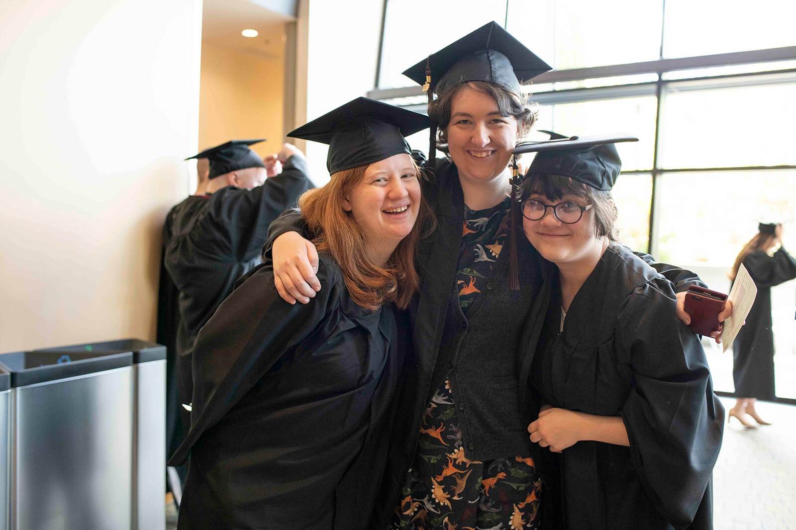 Three students in graduation robes and mortarboards embrace each other for a photo in a convention center hallway.