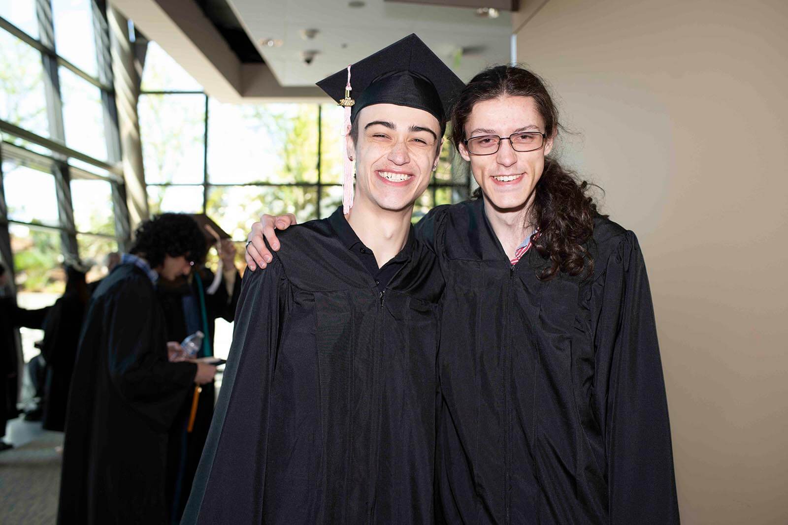 Two students in graduation garb smile for a photo in a convention center hallway.