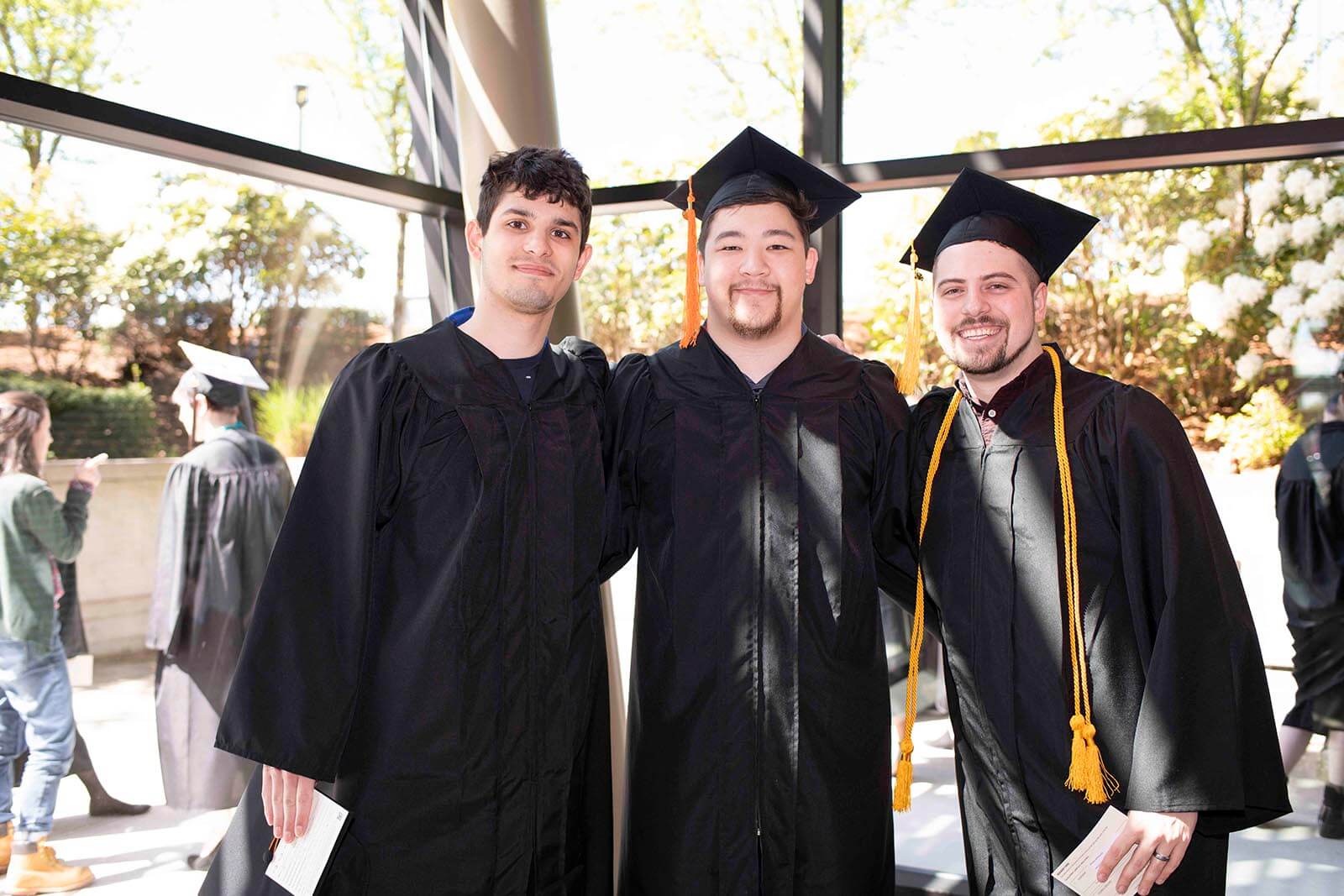 Three students in graduation robes and mortarboards pose for a photo in front of a floor-to-ceiling glass wall.