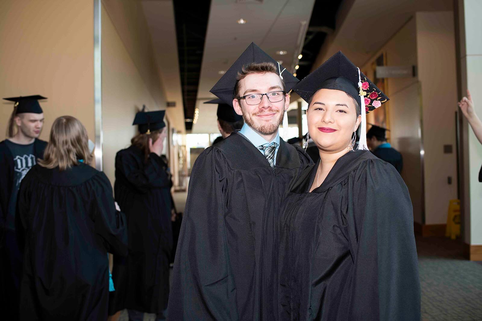 Two students in black graduation garb pose for a photo in a convention center hallway.