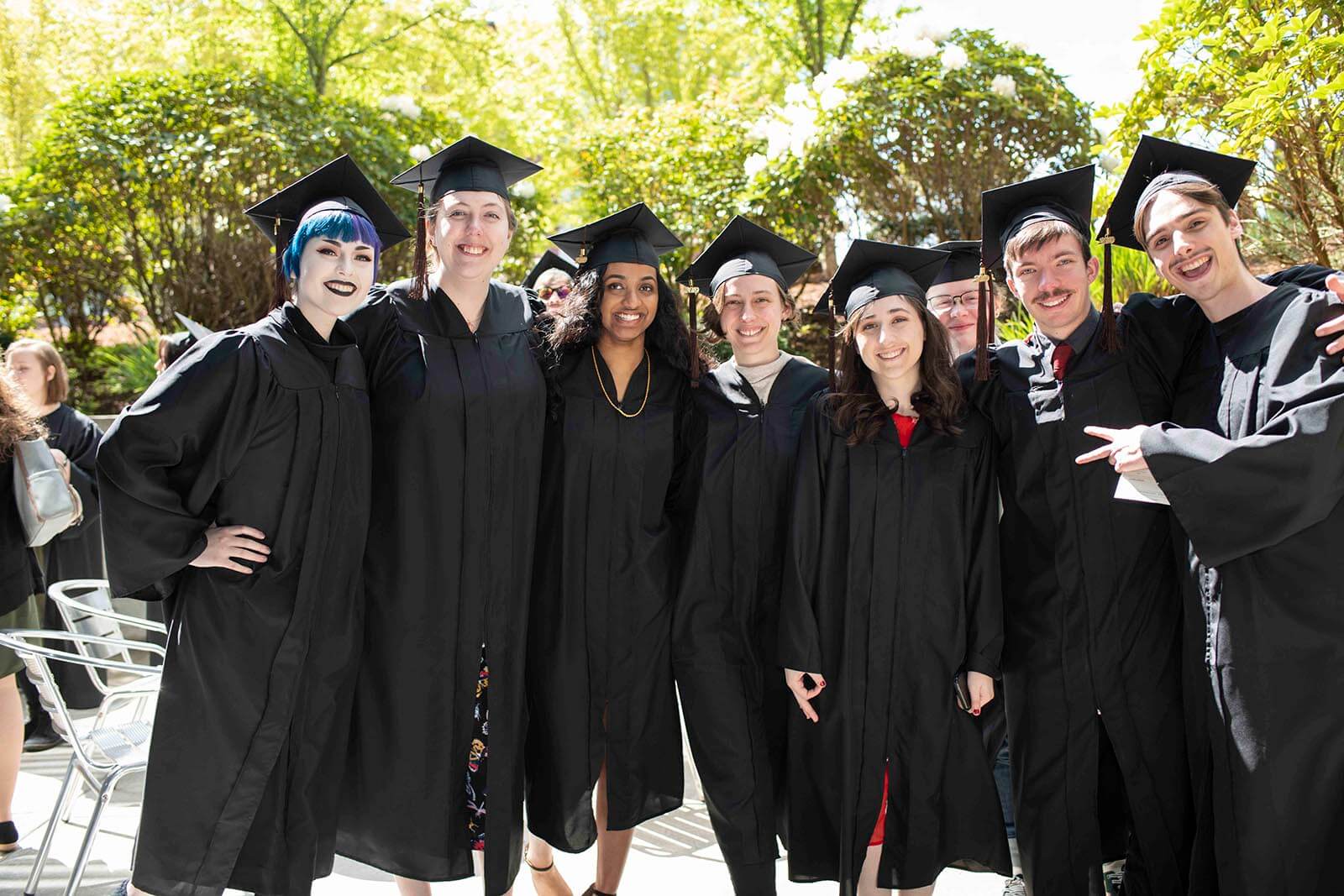 A group of students in graduation robes and mortarboards pose for a photo in a tree-lined courtyard.