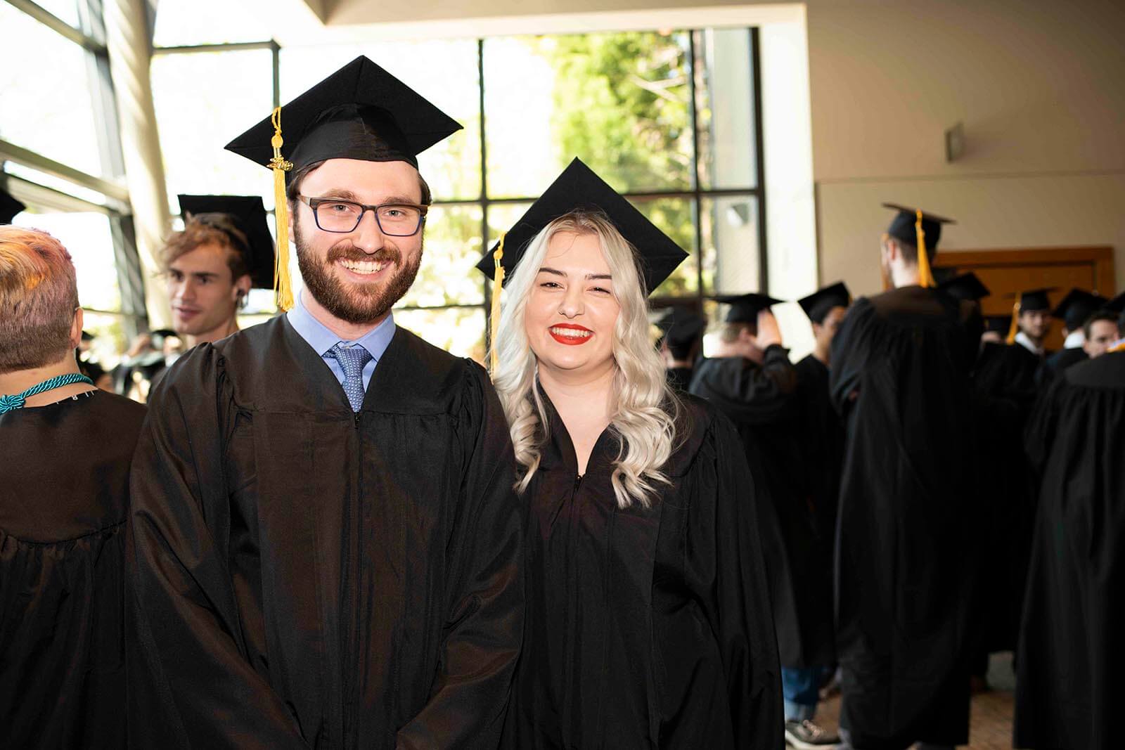 Two students in graduate robes and mortarboards smile for a photo in a convention center lobby.