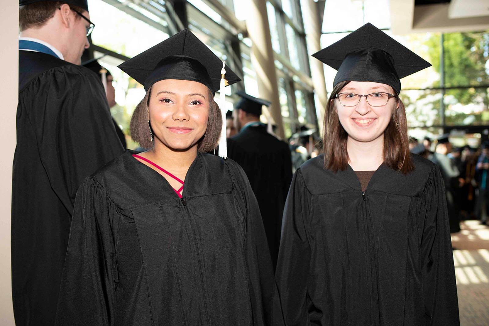 Two students in graduate garb smile for a photo in a convention center lobby area.