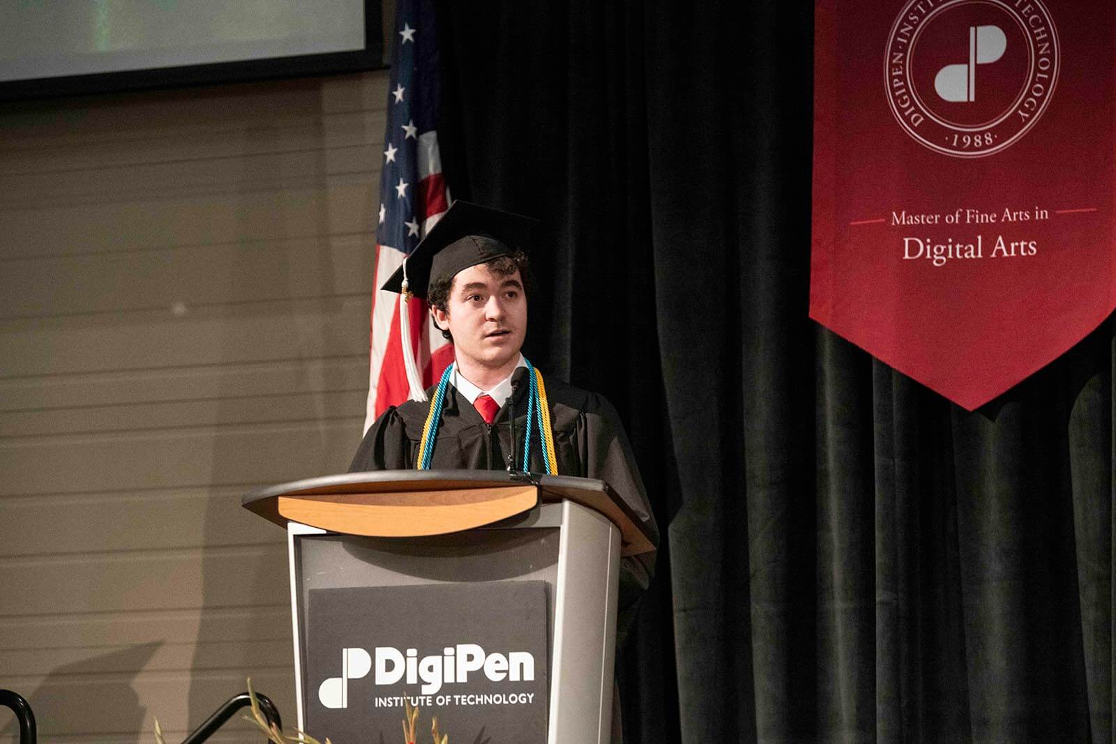 DigiPen commencement student speaker Christopher Onorati stands at a podium in graduate robe and mortarboard.