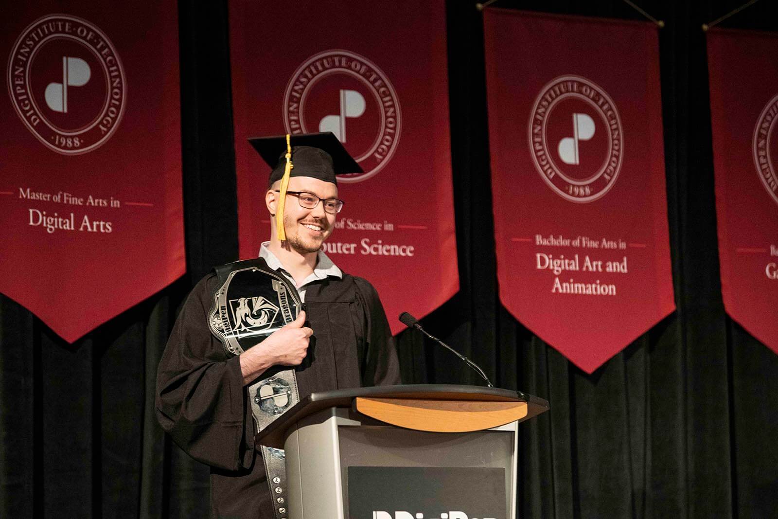 Benjamin Albertson-Gass stands at a podium in graduate garb holding a wrestling-style belt bearing the DigiPen logo.