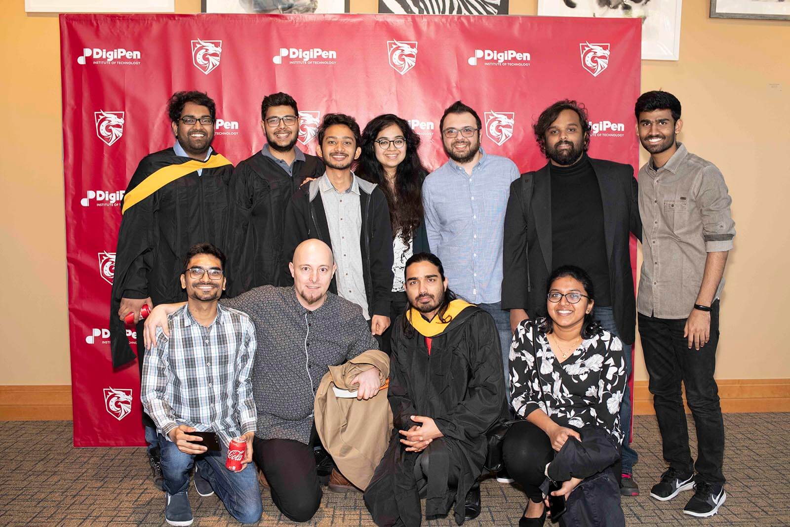 A group of people, some in graduation robes pose in front of a red backdrop bearing DigiPen's logo and name.