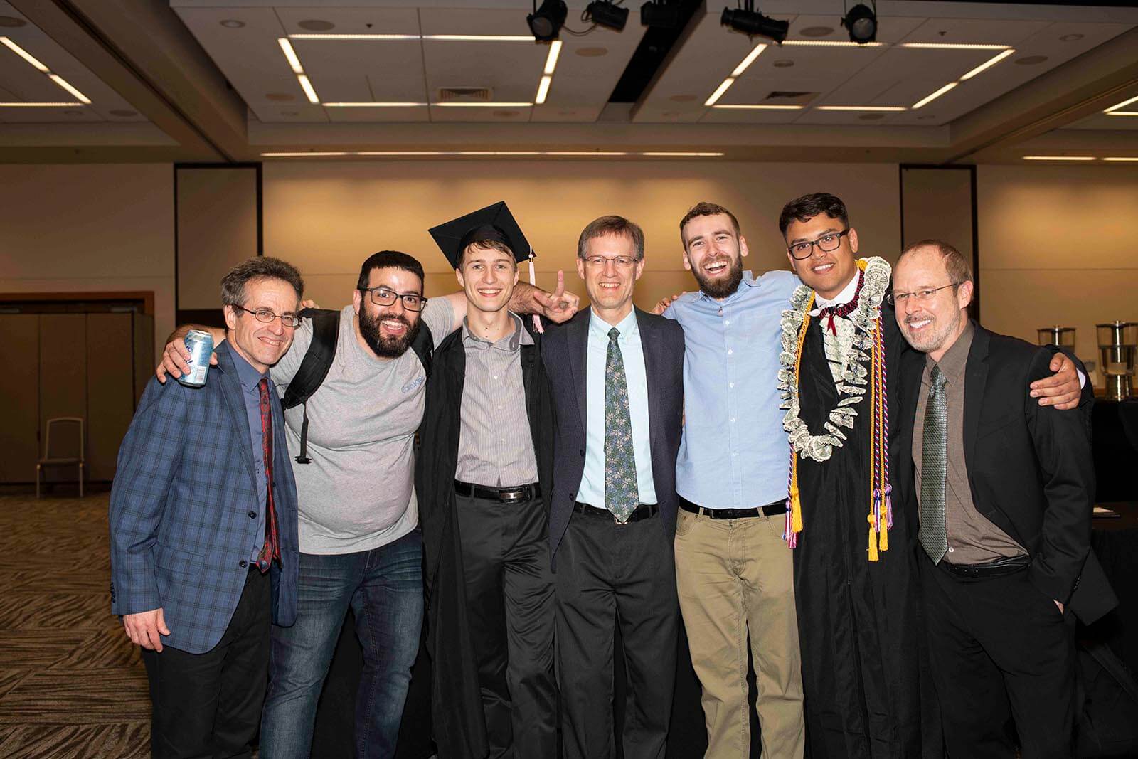 Students wearing graduate garb pose with DigiPen professors in a convention center meeting room.