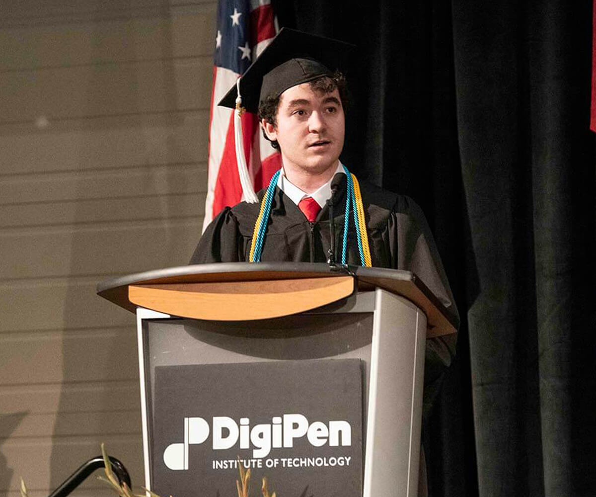 Chris Onorati speaks into a lectern microphone wearing his cap andgown at the 2019 DigiPen Commencement Ceremony. 