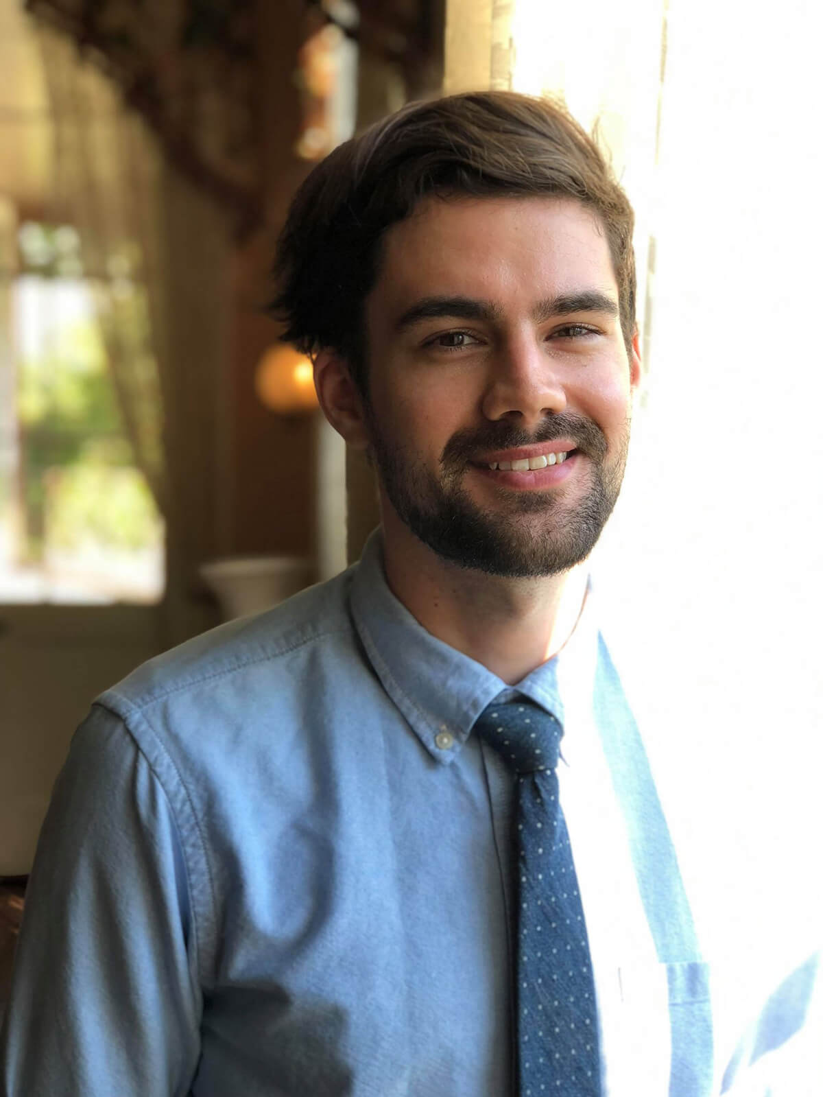 Man in a shirt and tie poses for a picture in a sunny window.