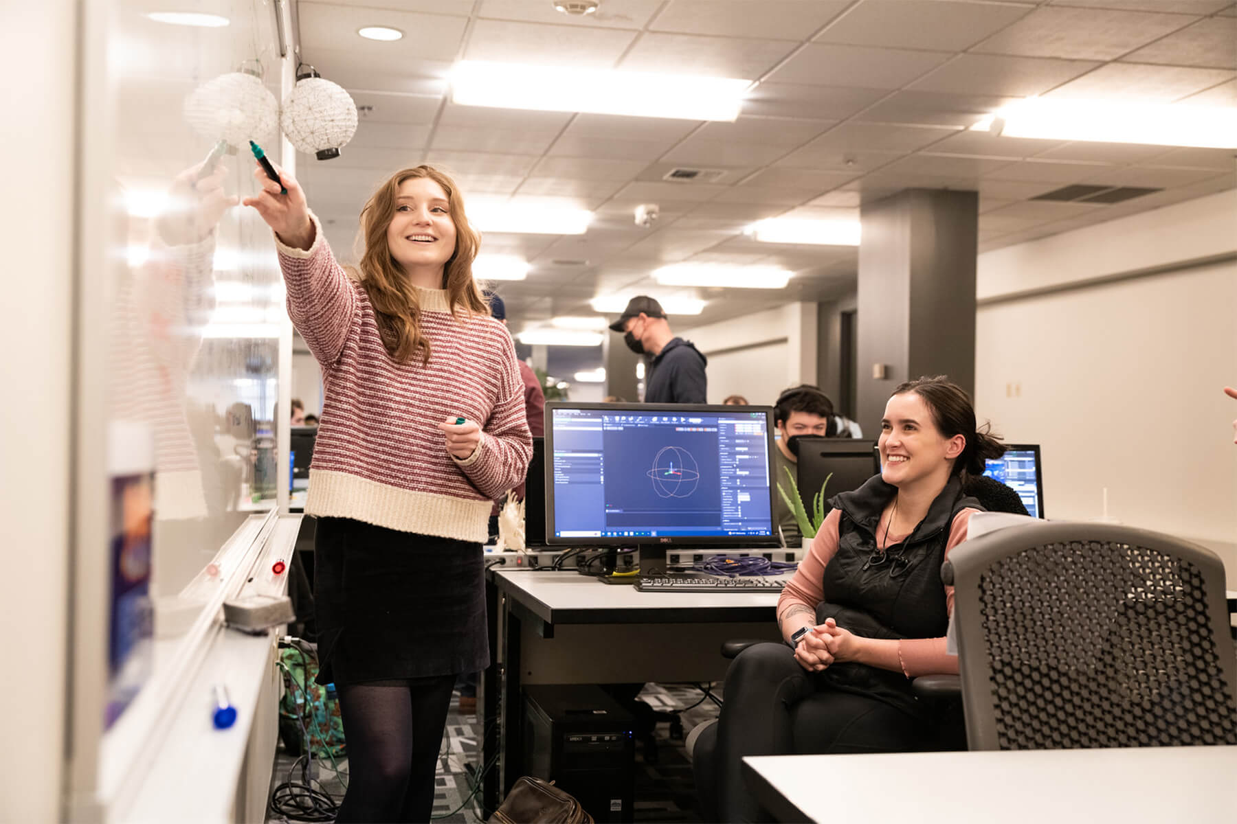 A DigiPen student points their marker at a whiteboard in front of a computer with another smiling student.