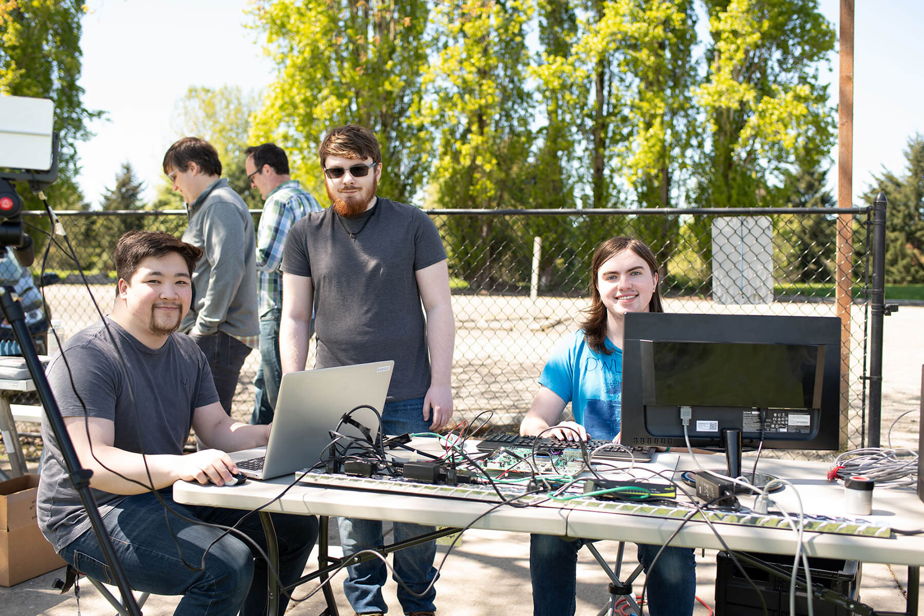 Three students pose in a park behind a computer setup that includes a laptop, monitor, radar system, and wires.