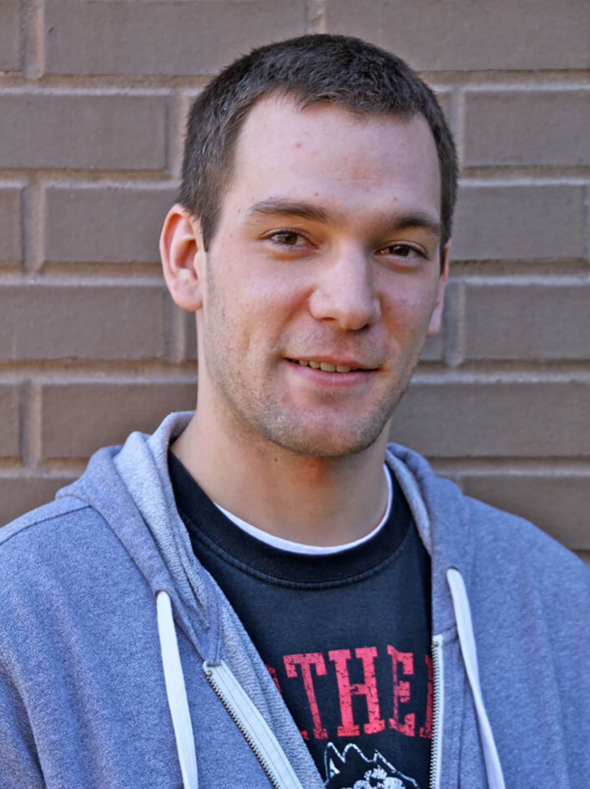 John Yednock smiles against a brick wall wearing a zip-up hoodie.