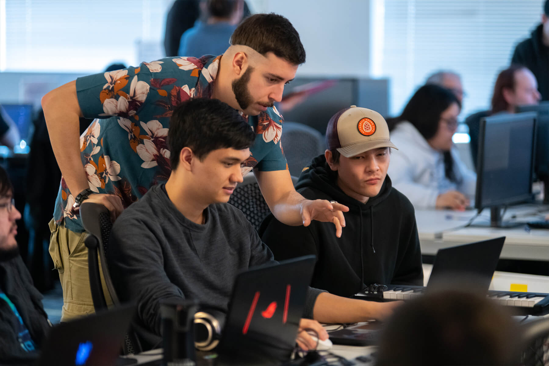 A student in a Hawaiian shirt gestures towards his teammate's computer screen as another team member looks on.