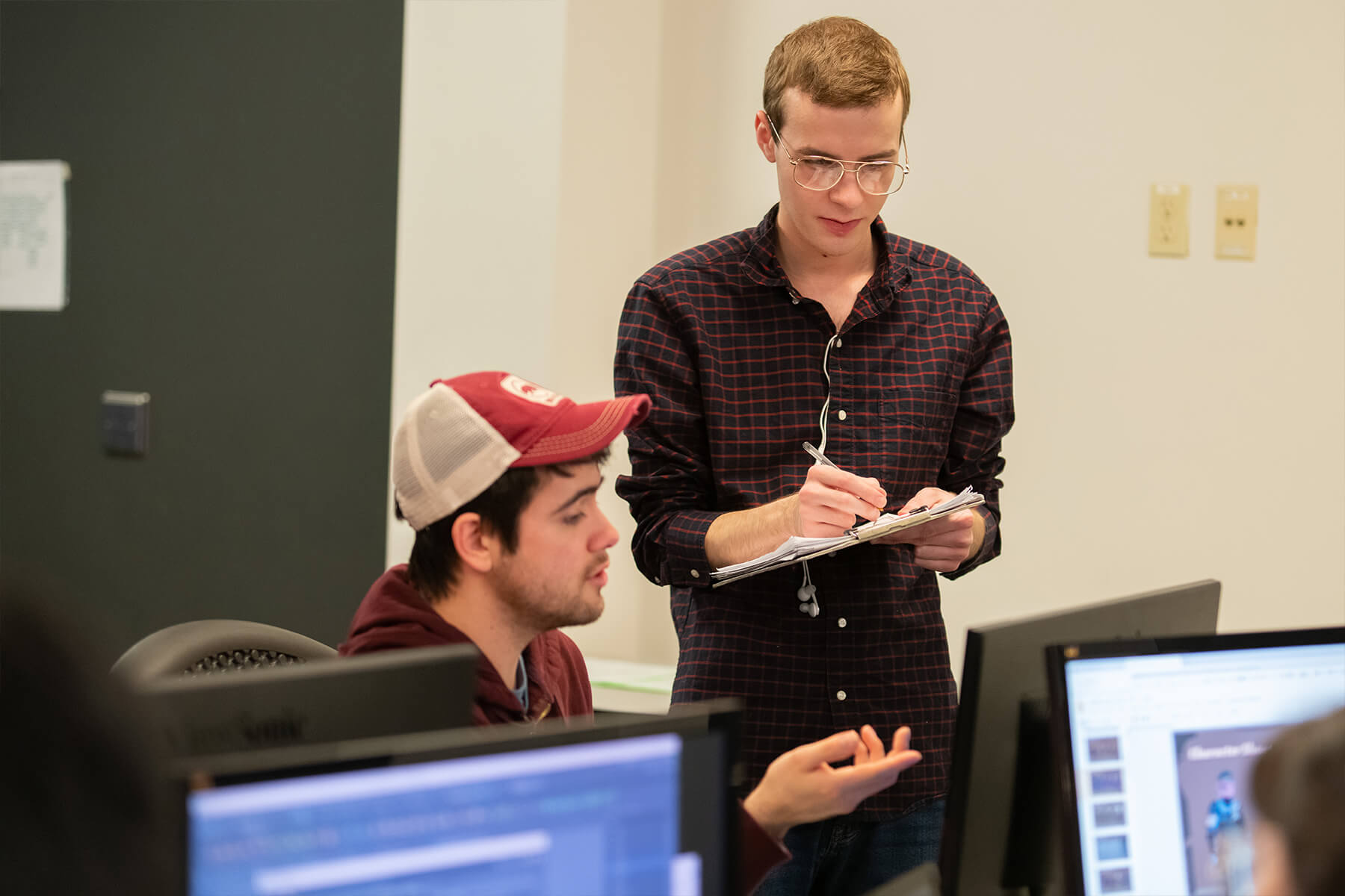 A student takes notes and sketches on a clipboard as their teammate talks and gestures at their screen. 