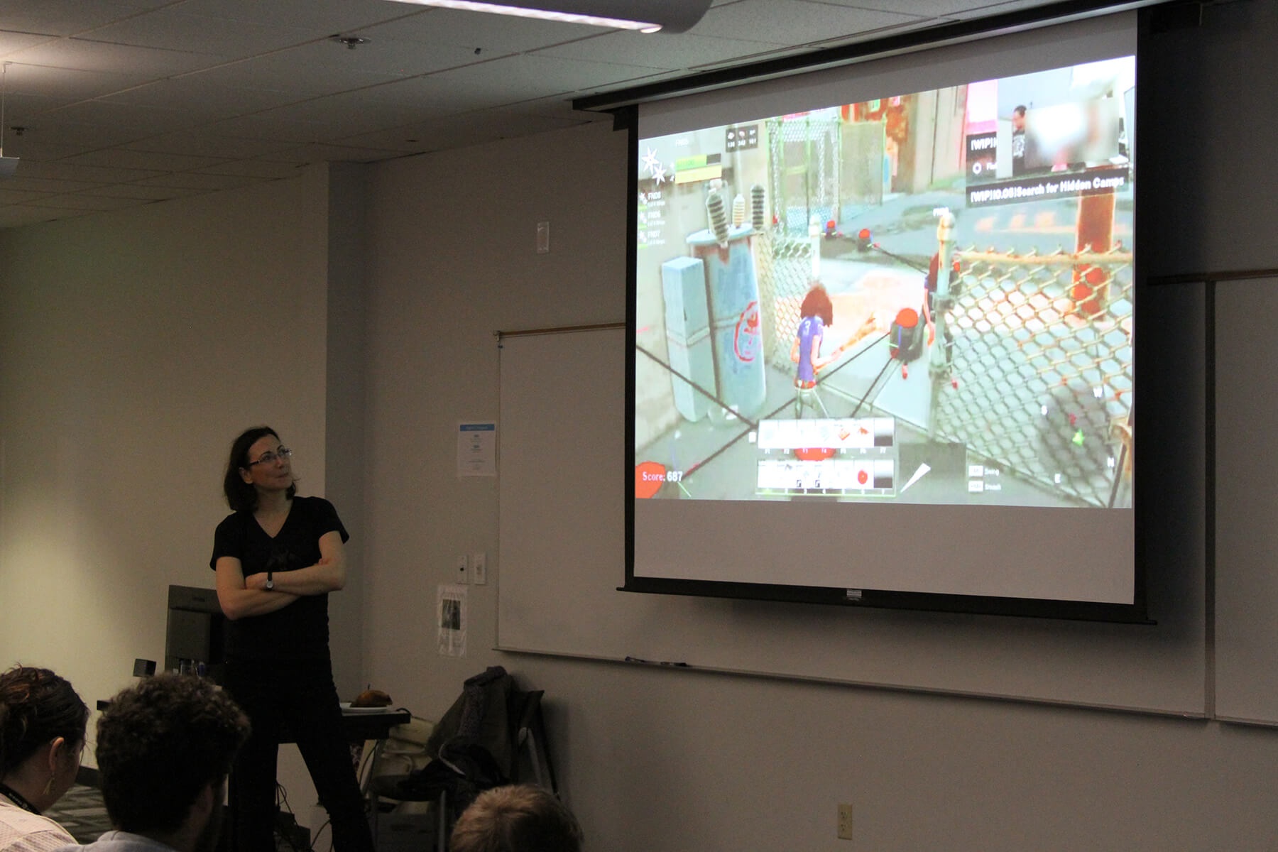 Dr. Hodent stands, arms crossed, in front of students during her lecture, scenes from a Fortnite play test being projected behind her. 