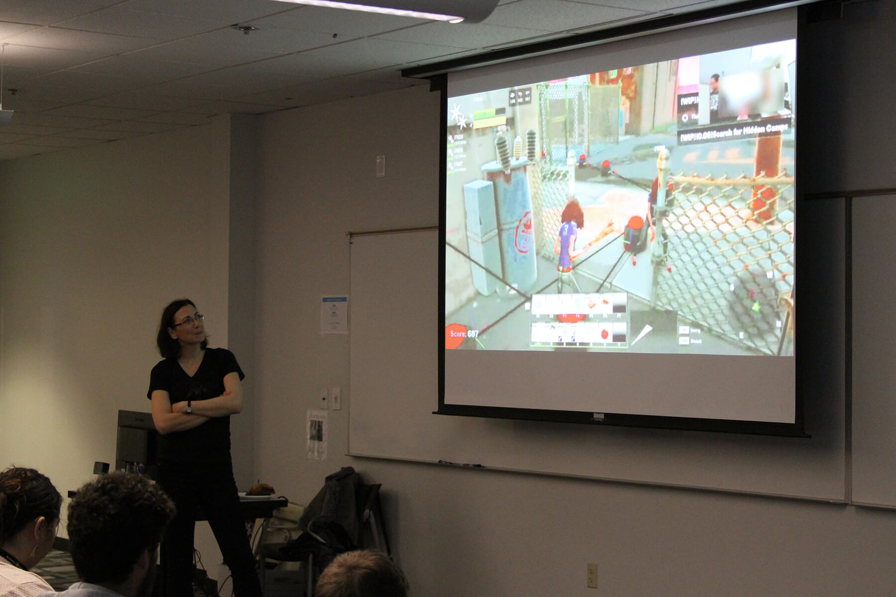 Dr. Hodent stands, arms crossed, in front of students during her lecture, scenes from a Fortnite play test being projected behind her.