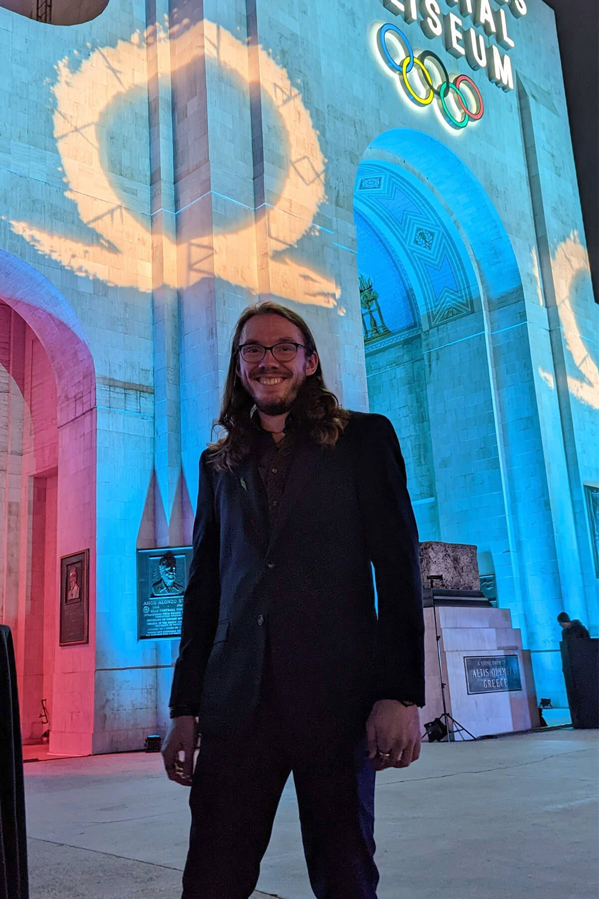 Josh Hammond poses in front of an Olympic coliseum with the God of War logo projected on it.