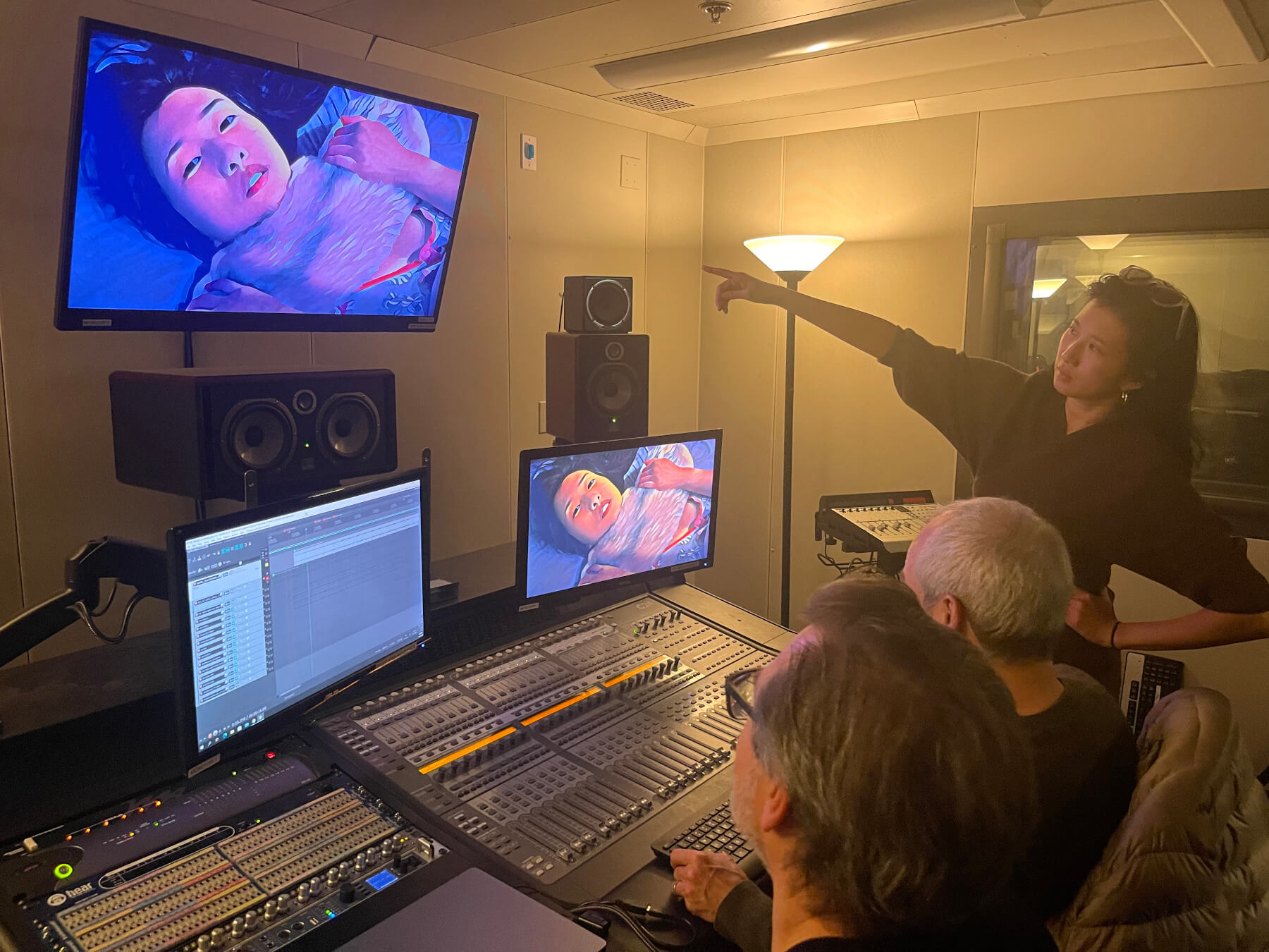 Chris Mosio and Lawrence Schwedler sit at studio sound board while Clare Chun stands and points at television monitor.