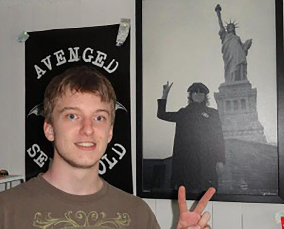 Joe Malmsten makes the peace sign while standing in front of a classic photo poster of John Lennon at the Statute of Liberty.