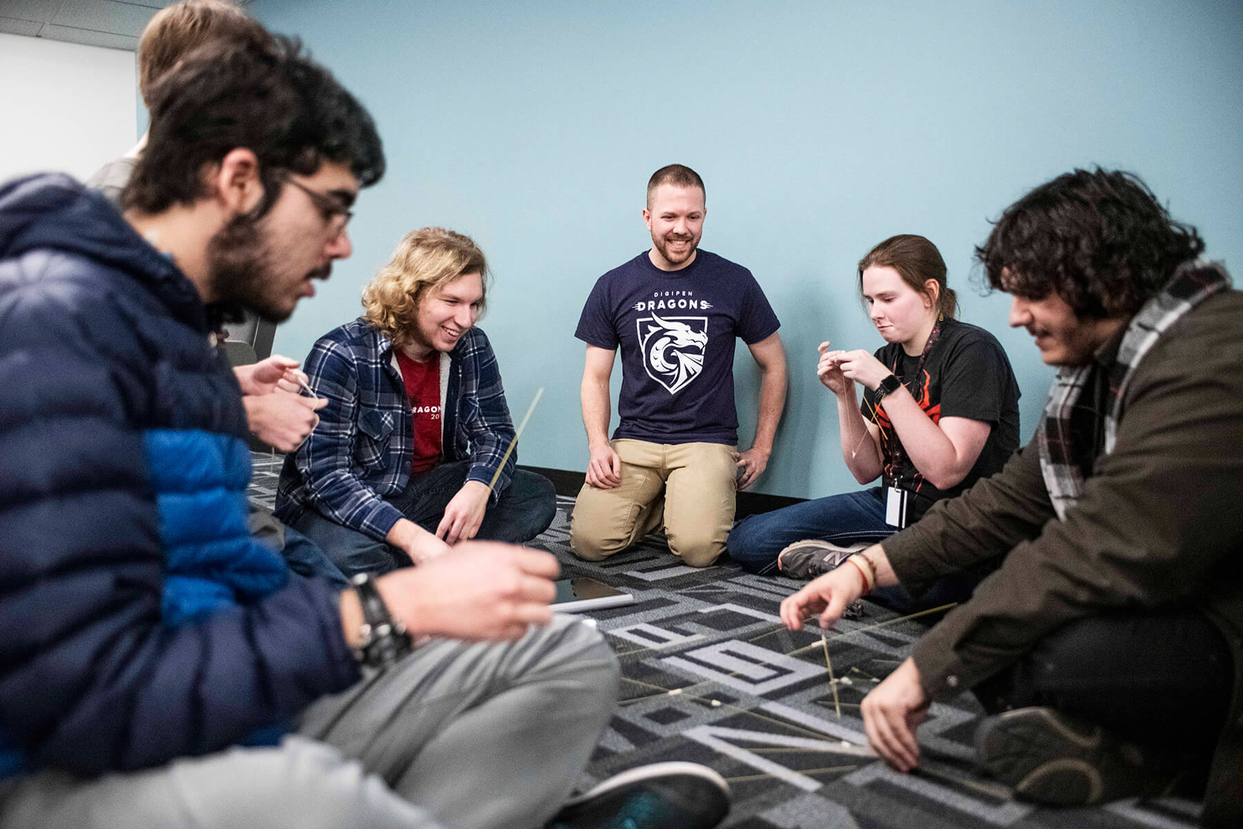 Five students and Justin Chambers sit in a semi-circle on classroom floor, building with pasta noodles and marshmallows.