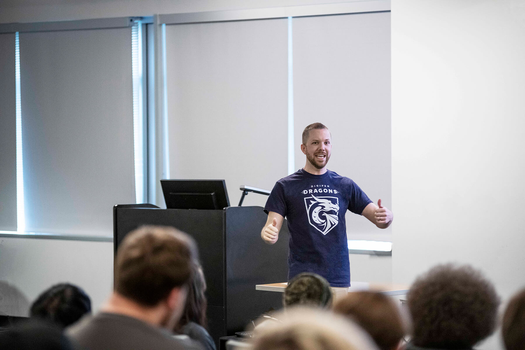 Justin Chambers stands near classroom lectern making thumbs up gesture with both hands while facing the seated students.